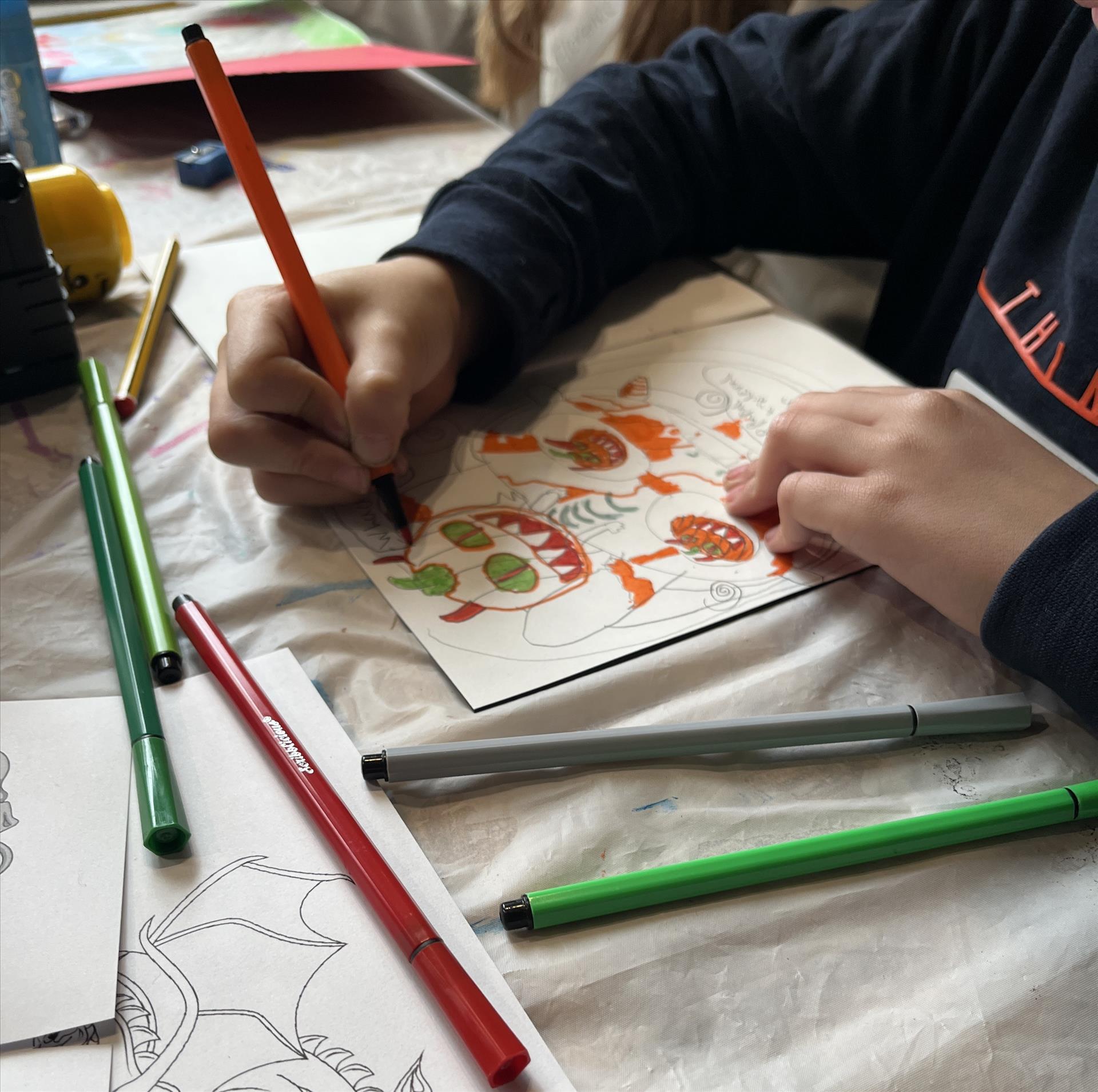 A table with craft materials on, with children's hands writing and drawing around the edge. 