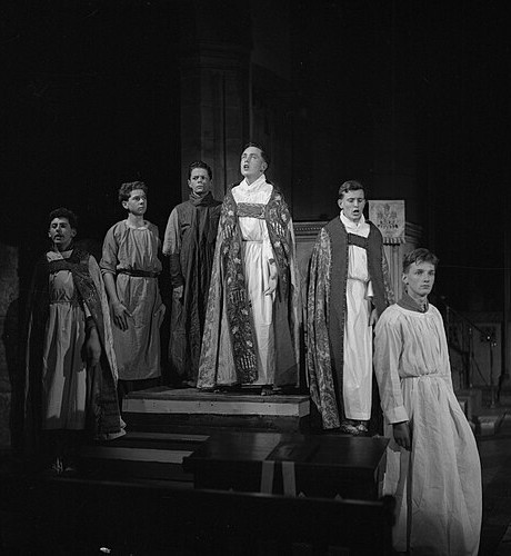 A black and white photograph of young people posed in the midst of performing a play in a cathedral. 