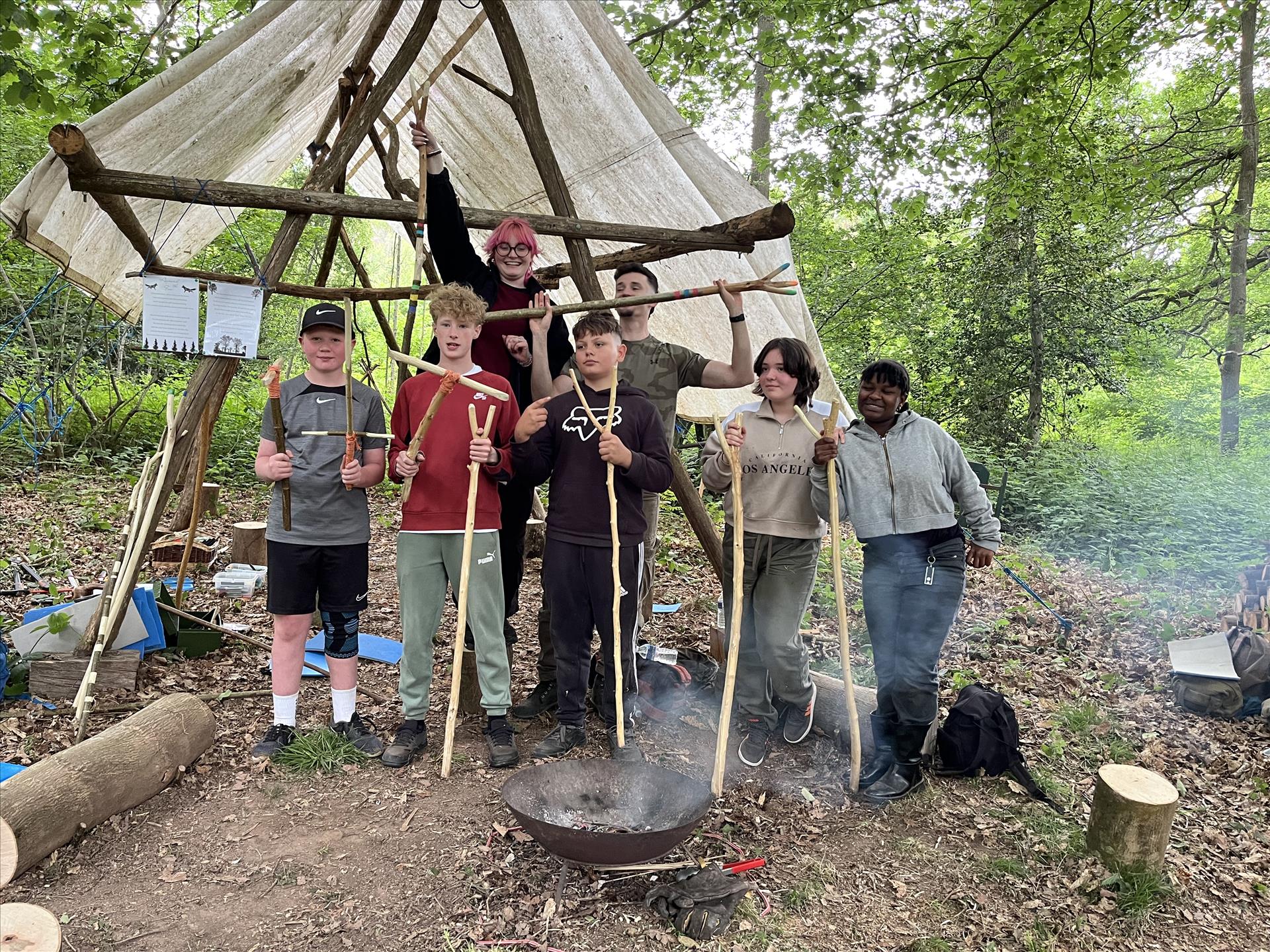 A group of young people standing in the woods, facing the camera. They are each holding a carved thumbstick.