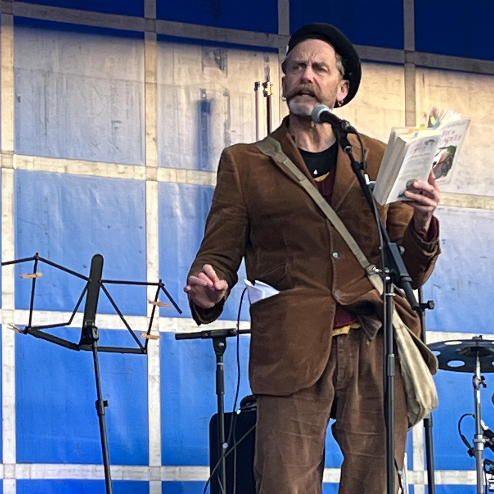 A man with a large moustache stood on a stage with blue lighting reading from a book. 