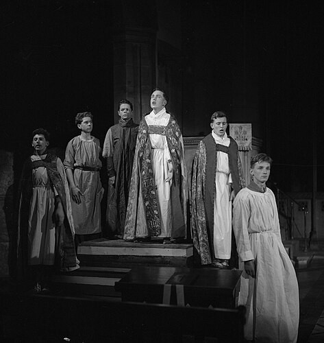 A black and white photograph of young people posed in the midst of performing a play in a cathedral. 