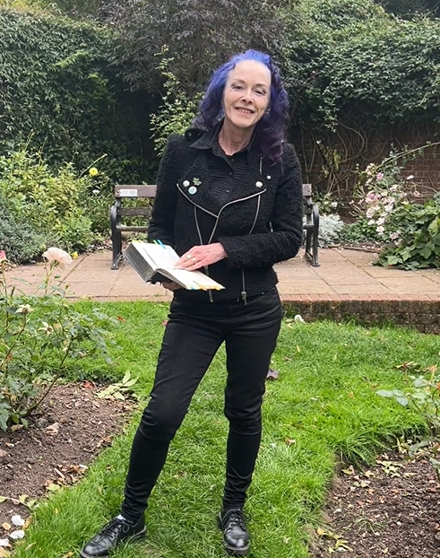 An image of a woman with curly blue hair and wearing black standing in a garden. 